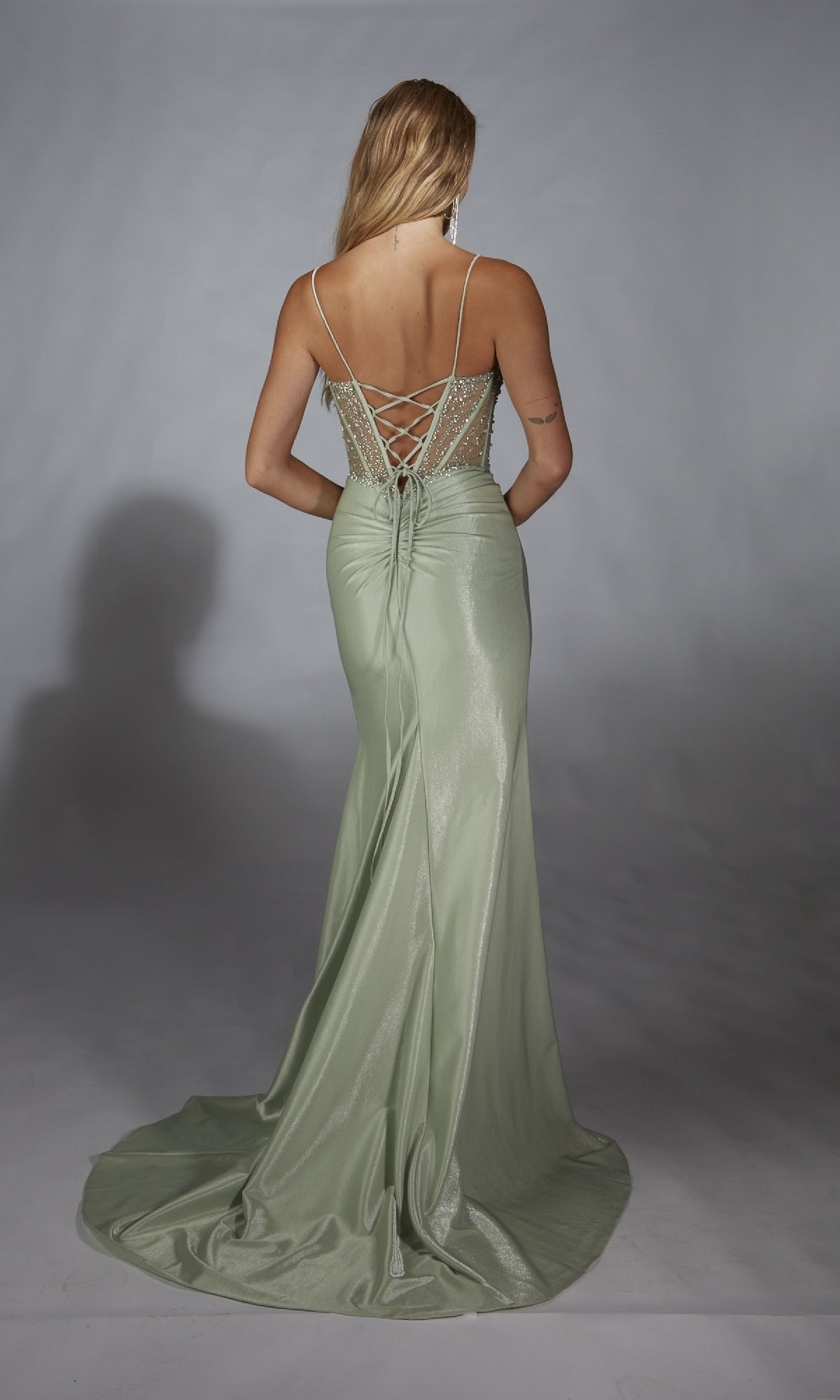 A woman stands facing away against a plain background, wearing the Alyce 61916—a light green, floor-length shimmer prom dress featuring a lace-up back.